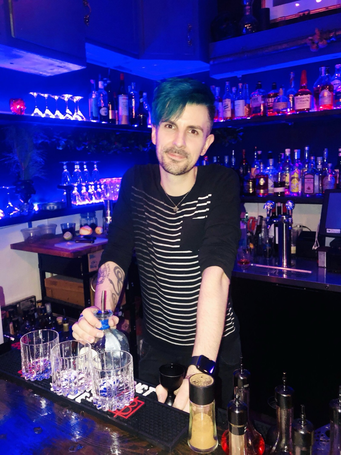 Kit standing behind a blue-lit bar with glassware set out in front of him