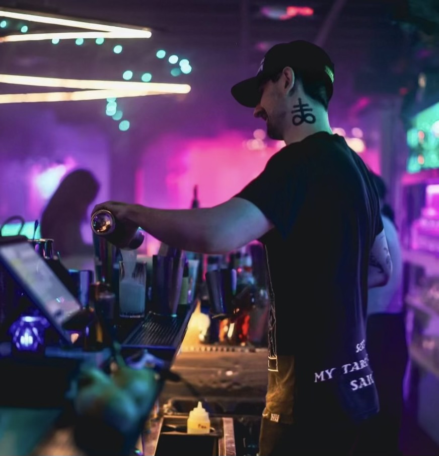Kit pouring a drink behind a bar under purple and teal lights